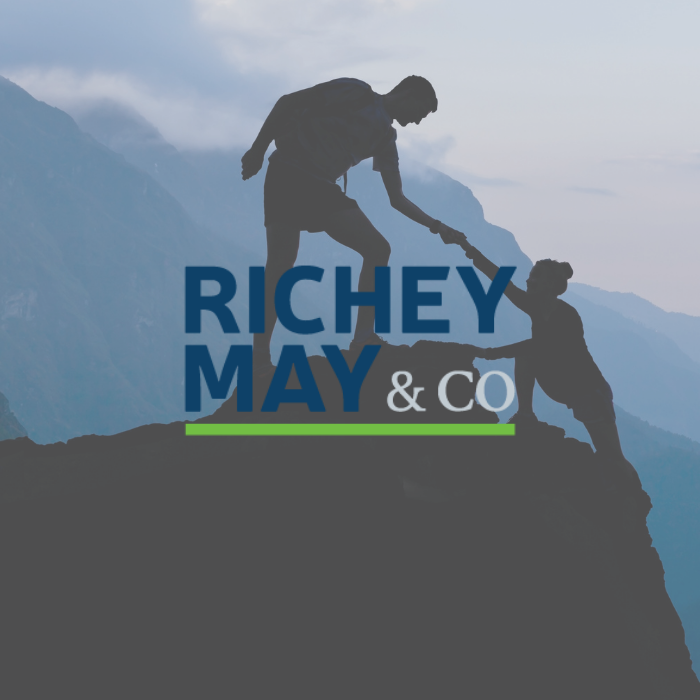 Richey May & Co title with background of a man helping his hiking partner up onto a rock
